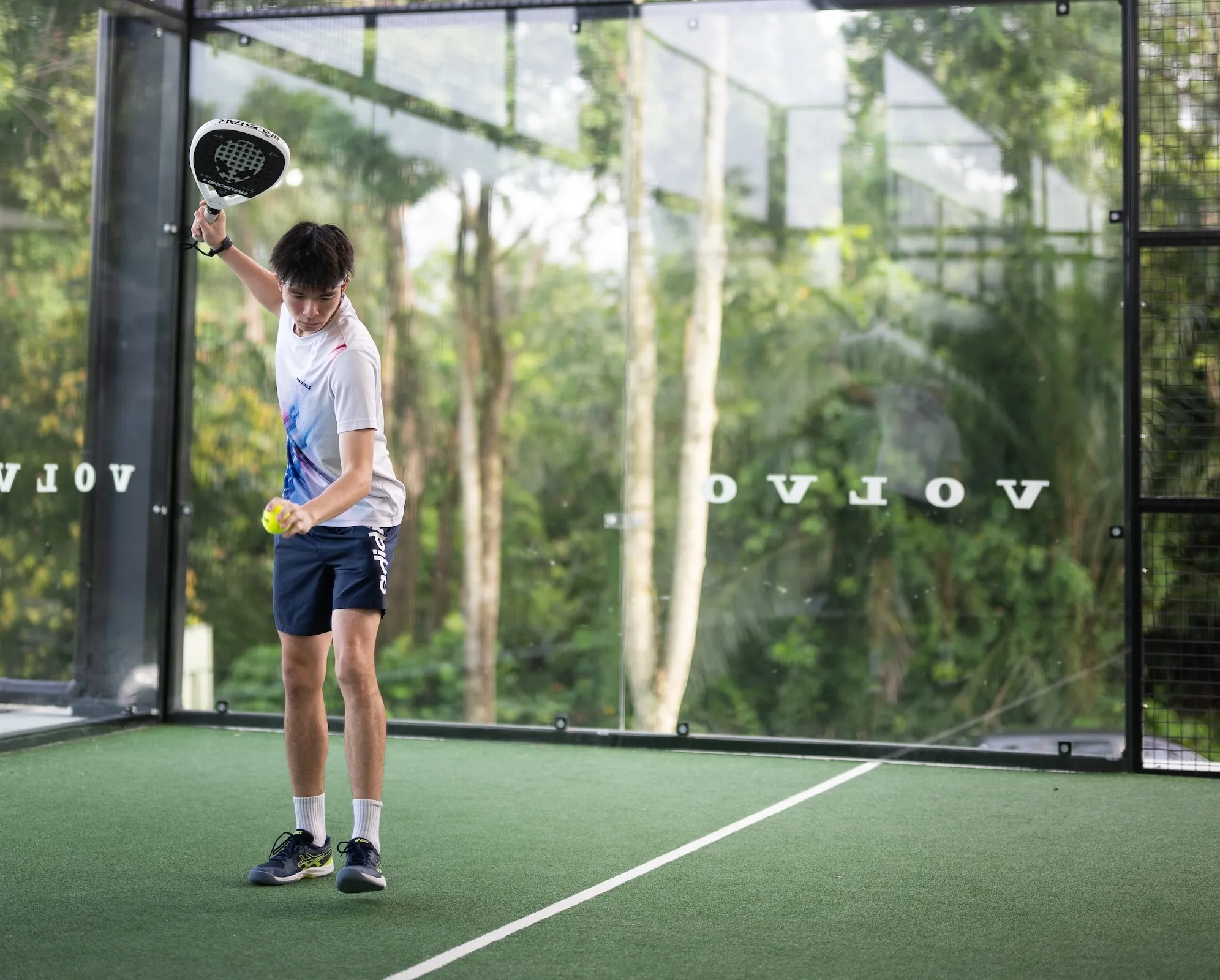 Aidan Yunus preparing to serve on a padel court, tropical forest visible through the glass